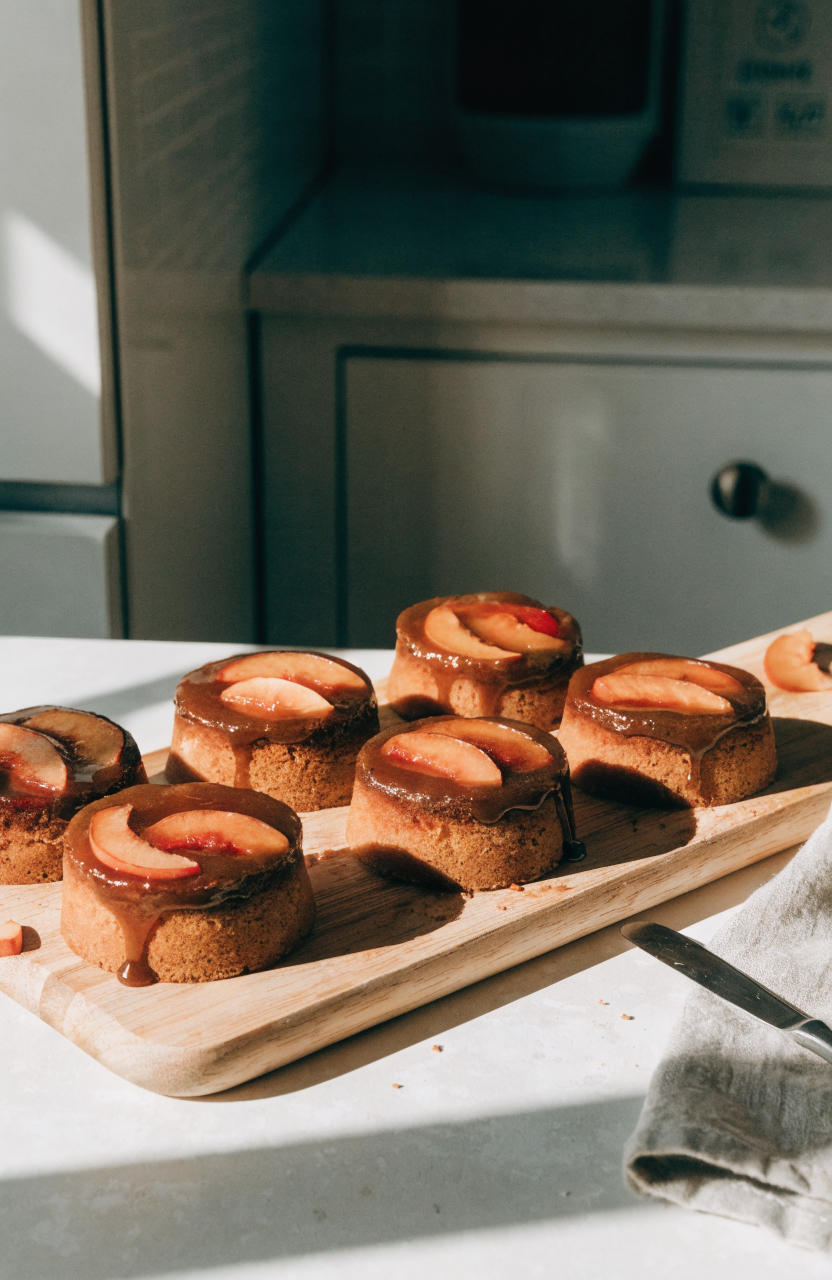 Six mini peach upside down cakes on a wooden board with glossy caramel tops and fresh peach slices